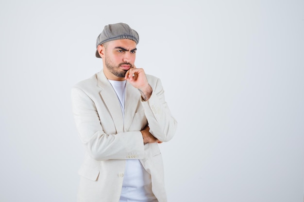 Jeune homme en t-shirt blanc, veste et casquette grise debout dans une pose de réflexion et l'air pensif