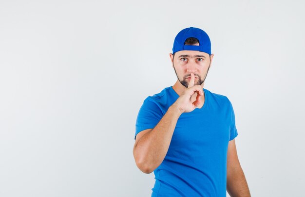 Jeune homme en t-shirt bleu et casquette montrant le geste de silence et à la prudence