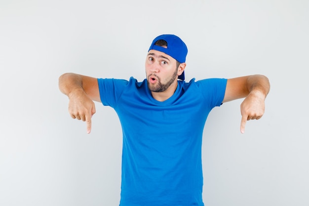 Photo gratuite jeune homme en t-shirt bleu et casquette pointant vers le bas et à la surprise