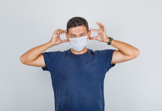 Jeune homme en t-shirt bleu foncé portant un masque médical et regardant attentivement, vue de face.