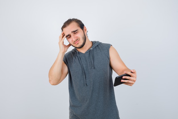 Jeune homme en t-shirt à capuche à la recherche de téléphone, tenant les doigts sur la tête et à la colère, vue de face.