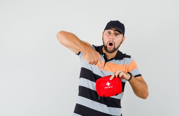 Jeune homme en t-shirt, casquette pointant sur la trousse de premiers soins et à la surprise, vue de face.