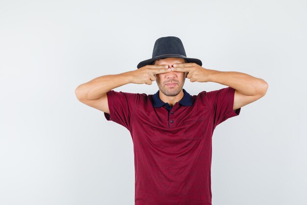 Jeune homme en t-shirt, chapeau couvrant les yeux avec les doigts, vue de face.