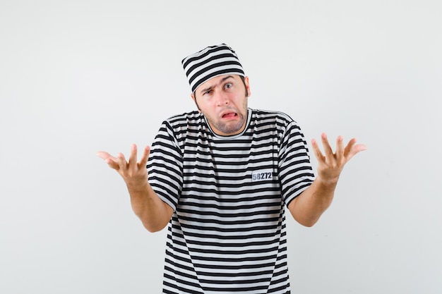 Jeune homme en t-shirt, chapeau étirant les mains dans un geste perplexe et à la drôle, vue de face.