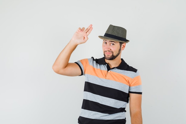 Jeune homme en t-shirt, chapeau faisant des gestes avec la main et les doigts et regardant confiant, vue de face.