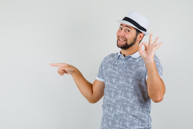Jeune homme en t-shirt, chapeau montrant un geste correct, pointant vers le côté et regardant gai, vue de face.