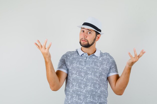 Jeune homme en t-shirt, chapeau montrant un geste impuissant et à la confusion, vue de face.