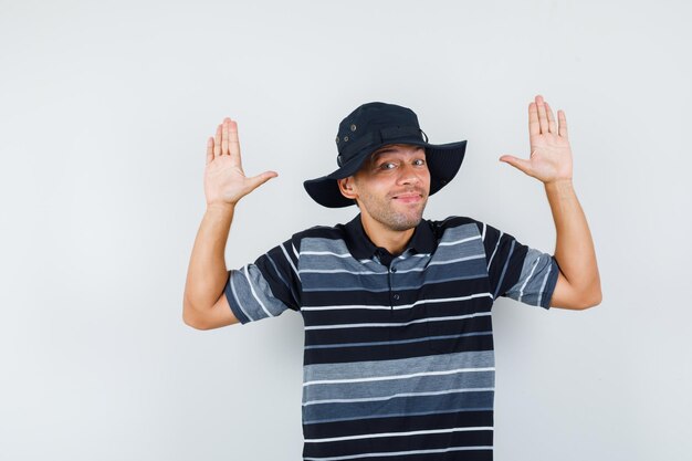 Jeune homme en t-shirt, chapeau montrant des paumes en geste d'abandon et l'air joyeux, vue de face.