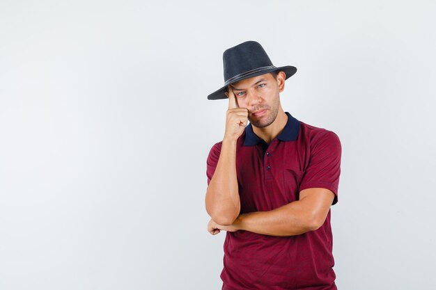 Jeune homme en t-shirt, chapeau penché sur la joue main levée et l'air pensif , vue de face.