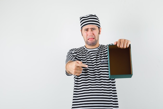 Jeune homme en t-shirt, chapeau pointant sur une boîte-cadeau vide et regardant vers le bas, vue de face.