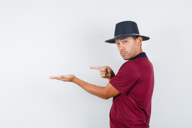 Jeune homme en t-shirt, chapeau pointant vers l'extérieur avec paume écartée et l'air confiant.