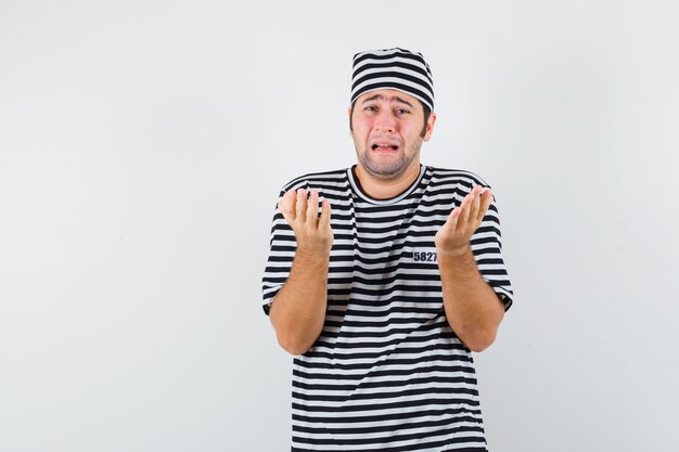 Jeune homme en t-shirt, chapeau priant en pleurant et regardant impuissant, vue de face.