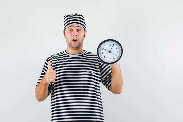 Jeune homme en t-shirt, chapeau tenant une horloge murale, montrant le pouce vers le haut et regardant excité, vue de face.