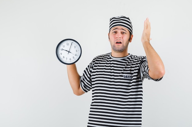 Jeune homme en t-shirt, chapeau tenant une horloge murale et regardant inquiet, vue de face.