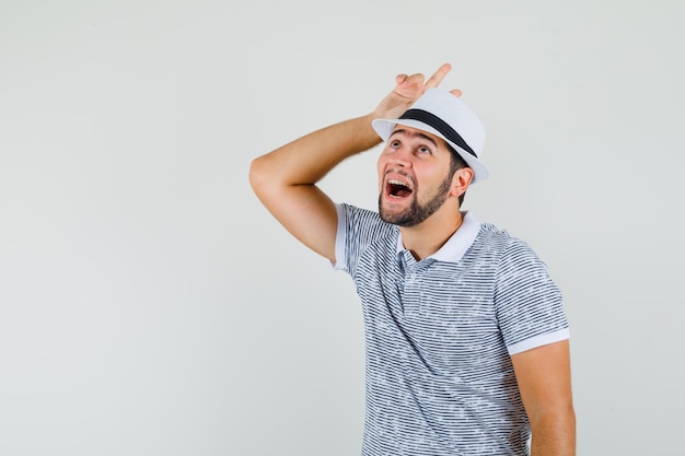 Jeune homme en t-shirt, chapeau tenant un signe v au-dessus de la tête comme des cornes et l'air drôle, vue de face.