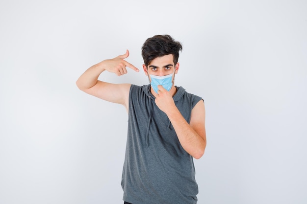 Jeune homme en t-shirt gris portant un masque tout en pointant sur lui-même et en ayant l'air sérieux