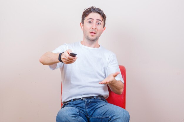 Jeune homme en t-shirt, jeans à l'aide de la télécommande alors qu'il était assis sur une chaise et l'air perplexe