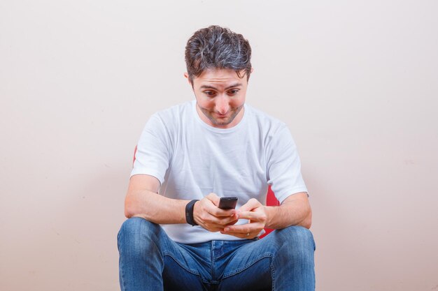 Jeune homme en t-shirt, jeans à l'aide de la télécommande assis sur une chaise