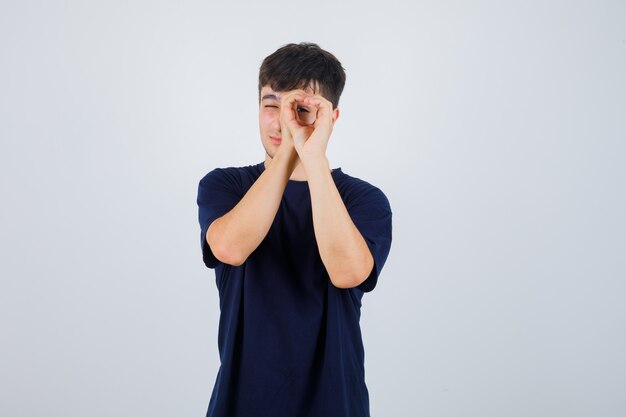 Jeune homme en t-shirt noir faisant semblant de jeter un coup d'œil à travers le trou avec ses mains et regardant curieux, vue de face.