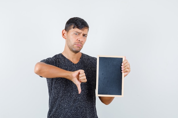 Jeune homme en t-shirt noir montrant le pouce vers le bas tout en tenant un carton noir et à l'insatisfaction, vue de face.