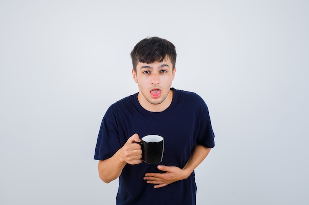 Jeune homme en t-shirt noir se sentir nauséeux tout en tenant une tasse de thé et à la recherche de mal, vue de face.