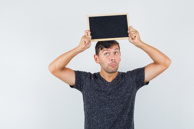 Jeune homme en t-shirt noir soulevant du carton noir sur sa tête et à la vue réfléchie, de face.