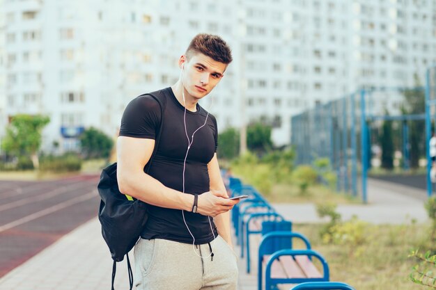 Jeune homme en T-shirt noir sport et pantalon de sport gris est debout sur fond de ville et de stade. Il écoute de la musique avec des écouteurs, tient un sac de sport et regarde la caméra.