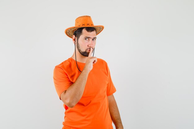 Jeune homme en t-shirt orange, chapeau montrant un geste de silence et regardant attentivement, vue de face.