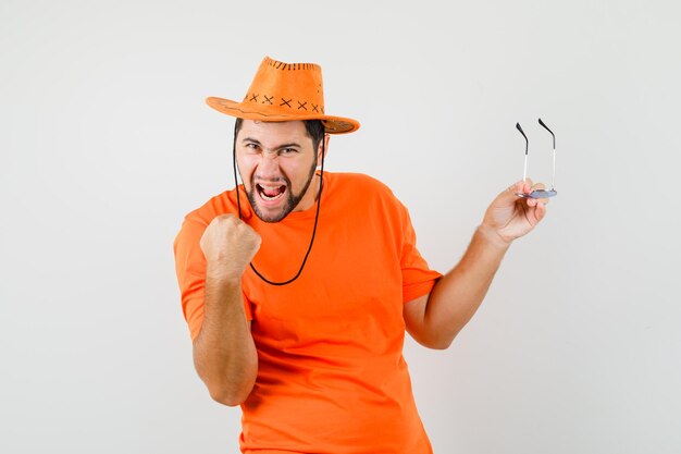 Jeune homme en t-shirt orange, chapeau tenant des lunettes avec le geste du gagnant et l'air heureux, vue de face.