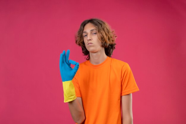 Jeune homme en t-shirt orange portant des gants en caoutchouc à la confiance de faire signe ok debout sur fond rose
