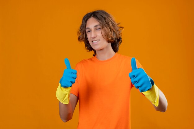 Jeune homme en t-shirt orange portant des gants en caoutchouc à la confiance en souriant joyeusement montrant les pouces vers le haut avec les deux mains debout sur fond jaune