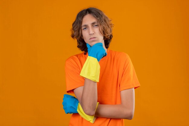 Jeune homme en t-shirt orange portant des gants en caoutchouc debout avec la main sur le menton regardant la caméra avec une expression pensive pensant sur fond jaune