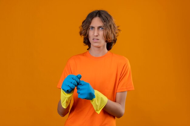 Jeune homme en t-shirt orange portant des gants en caoutchouc à la position incertaine et confuse sur fond jaune