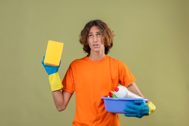 Jeune homme en t-shirt orange portant des gants en caoutchouc tenant le bassin avec des outils de nettoyage et une éponge à la recherche avec une expression triste sur le visage debout sur fond vert