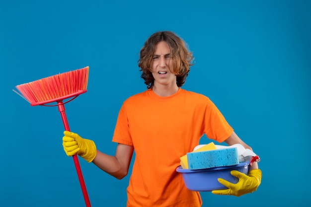 Photo gratuite jeune homme en t-shirt orange portant des gants en caoutchouc tenant le bassin avec des outils de nettoyage et une vadrouille à l'énervé debout sur fond bleu