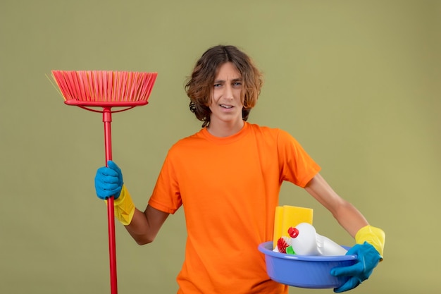 Jeune homme en t-shirt orange portant des gants en caoutchouc tenant bassin avec des outils de nettoyage et une vadrouille regardant la caméra confus et incertain debout sur fond vert