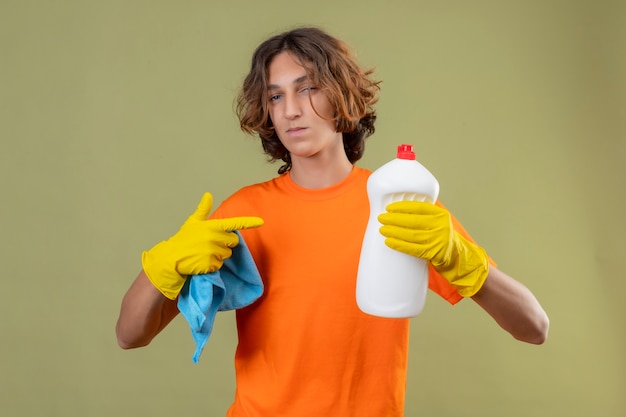 Jeune homme en t-shirt orange portant des gants en caoutchouc tenant une bouteille de produits de nettoyage pointant avec le doigt dessus à la confiance debout sur fond vert