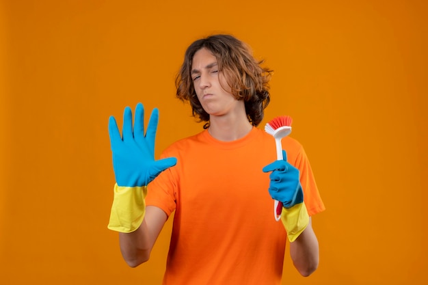 Jeune homme en t-shirt orange portant des gants en caoutchouc tenant une brosse à récurer debout avec la main ouverte faisant signe d'arrêt geste de défense sur fond jaune