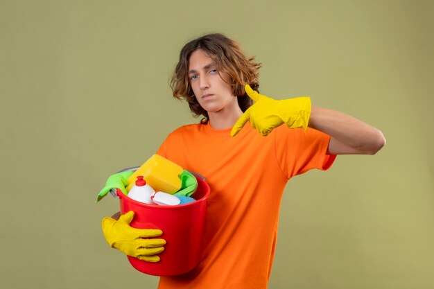 Jeune homme en t-shirt orange portant des gants en caoutchouc tenant un seau avec des outils de nettoyage pointant avec le doigt dessus à la caméra t avec une expression faciale confiante et sérieuse debout sur gre
