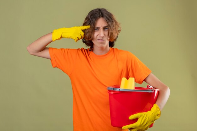 Jeune homme en t-shirt orange portant des gants en caoutchouc tenant seau avec des outils de nettoyage pointant le temple avec une expression sceptique sur le visage debout sur fond vert