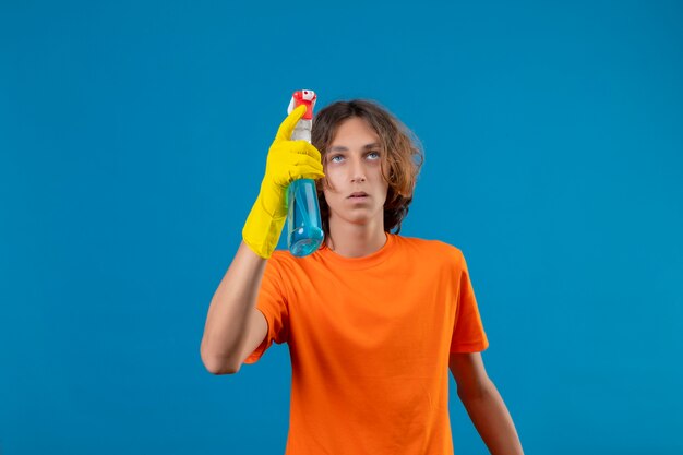 Jeune homme en t-shirt orange portant des gants en caoutchouc tenant un spray de nettoyage à la recherche avec une expression pensive sur le visage debout sur fond bleu