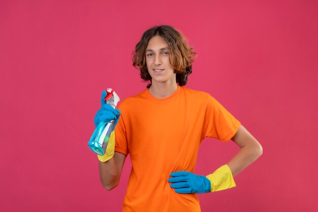 Jeune homme en t-shirt orange portant des gants en caoutchouc tenant un spray de nettoyage regardant la caméra avec un sourire confiant debout sur fond rose