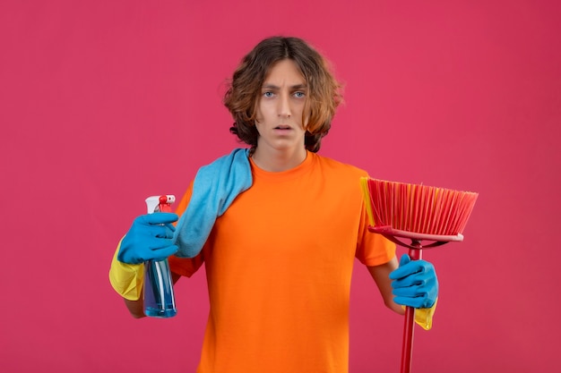 Jeune homme en t-shirt orange portant des gants en caoutchouc tenant une vadrouille et un spray de nettoyage regardant la caméra nerveuse et anxieuse debout sur fond rose