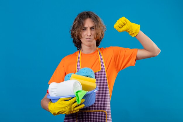 Jeune homme en t-shirt orange portant un tablier et des gants en caoutchouc tenant bassin avec des outils de nettoyage levant le poing avec un regard confiant se réjouissant de son succès et de sa victoire debout sur bac bleu