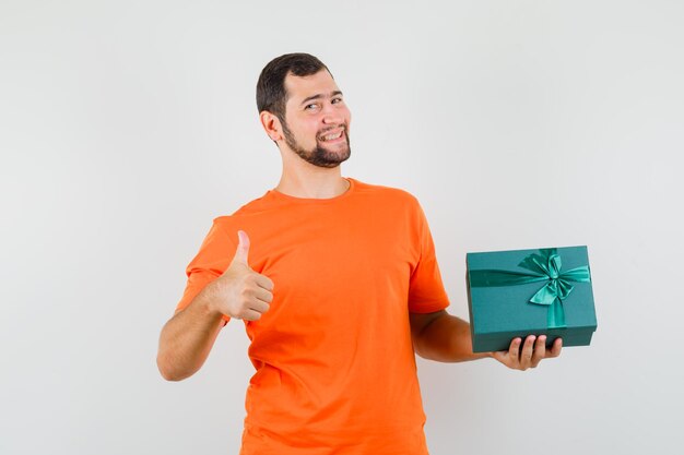 Jeune homme en t-shirt orange tenant une boîte présente avec le pouce vers le haut et à la gaieté, vue de face.