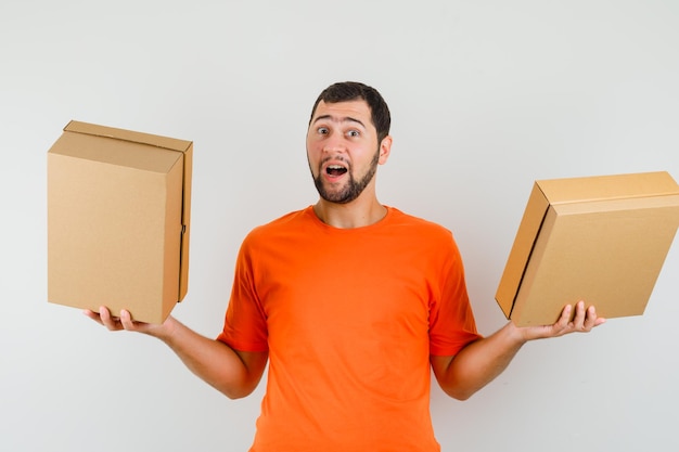 Jeune homme en t-shirt orange tenant des boîtes en carton et l'air optimiste, vue de face.