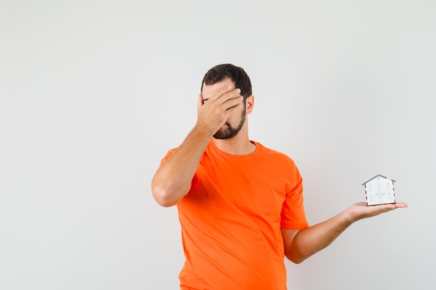 Jeune homme en t-shirt orange tenant un modèle de maison avec la main sur le visage, vue de face.