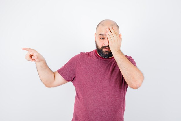 Jeune homme en t-shirt rose couvrant son seul œil tout en pointant de côté et l'air bizarre, vue de face.