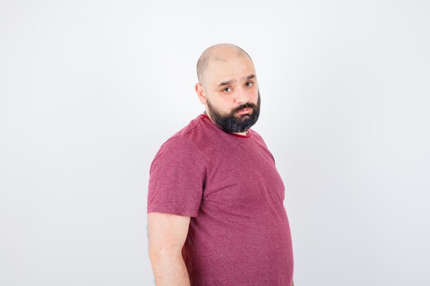 Jeune homme en t-shirt rose debout tout droit, regardant par-dessus l'épaule et se présentant à la caméra et ayant l'air sérieux, vue de face.