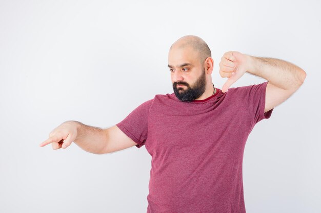 Jeune homme en t-shirt rose montrant le pouce vers le bas tout en pointant de côté et l'air nerveux, vue de face.